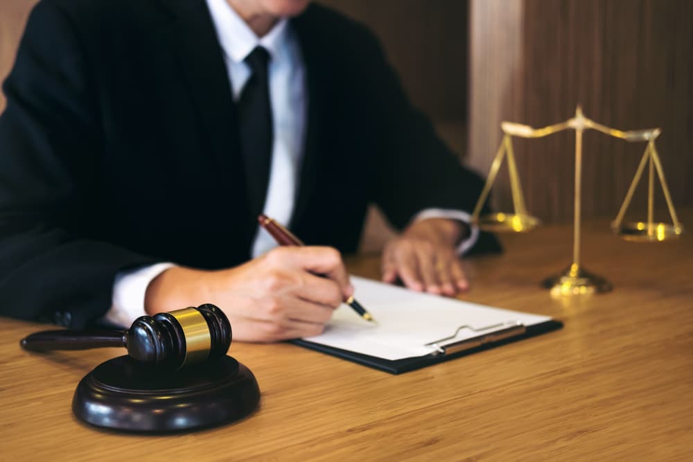 A judge's gavel sits next to justice scales as a businessman or lawyer in a suit works on legal documents. This scene represents the concept of law, legal advice, and justice.