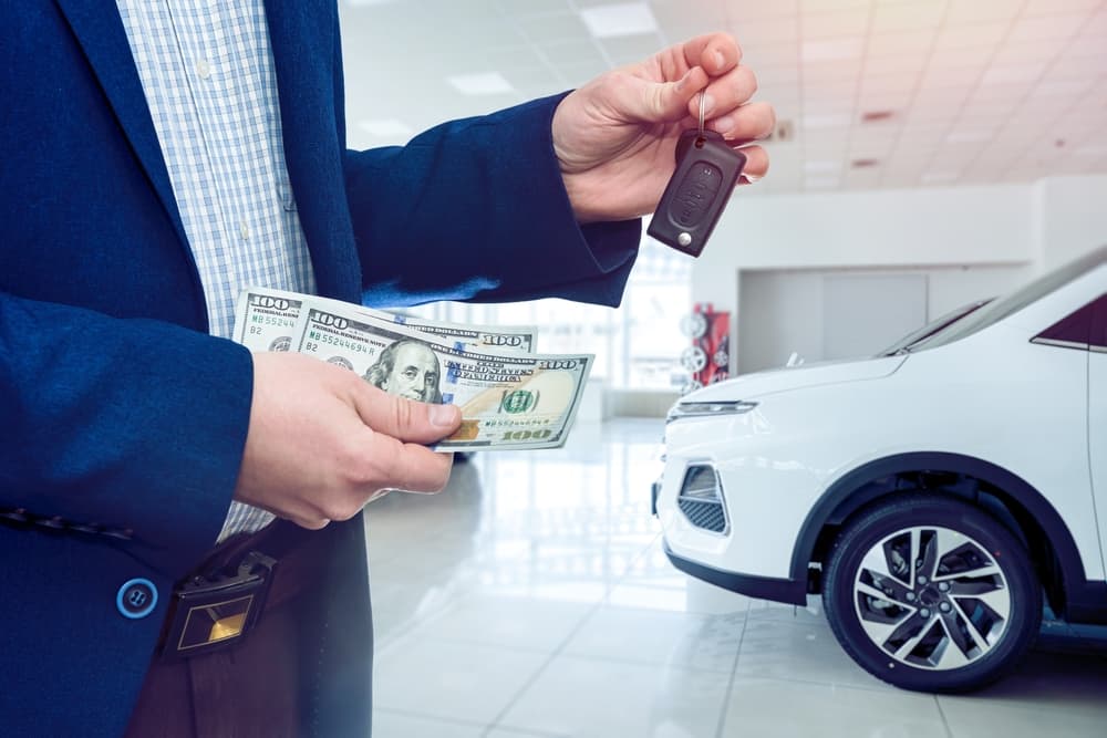 A male hand holding dollar banknotes near a car in a showroom, representing the concept of a car loan and purchasing a vehicle.







