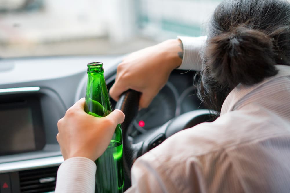 A driver holding a green bottle behind the wheel, representing the consequences of DWI offenses.