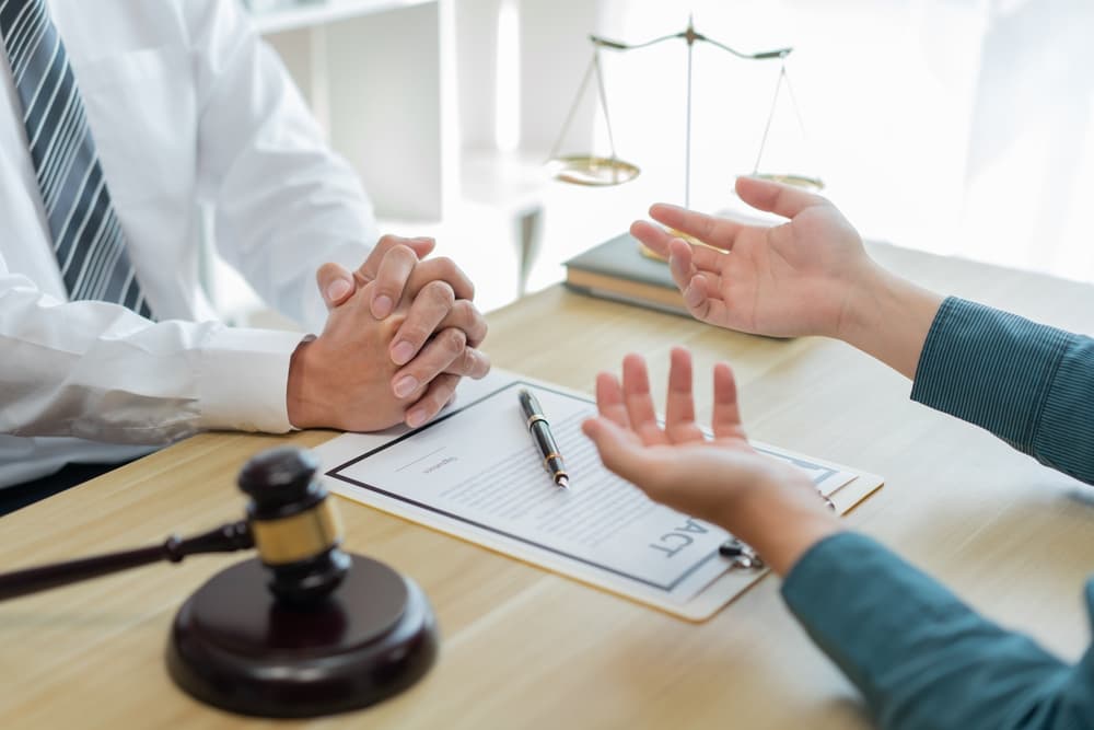 A male client sits across from a prosecutor in a professional office setting, gesturing with his hand as he explains a point of concern. The prosecutor, dressed formally, listens attentively while reviewing a document, possibly a contract, spread out on the desk. The desk is cluttered with legal papers, a laptop, and a pen, emphasizing the consultancy setting. The atmosphere is serious and focused.