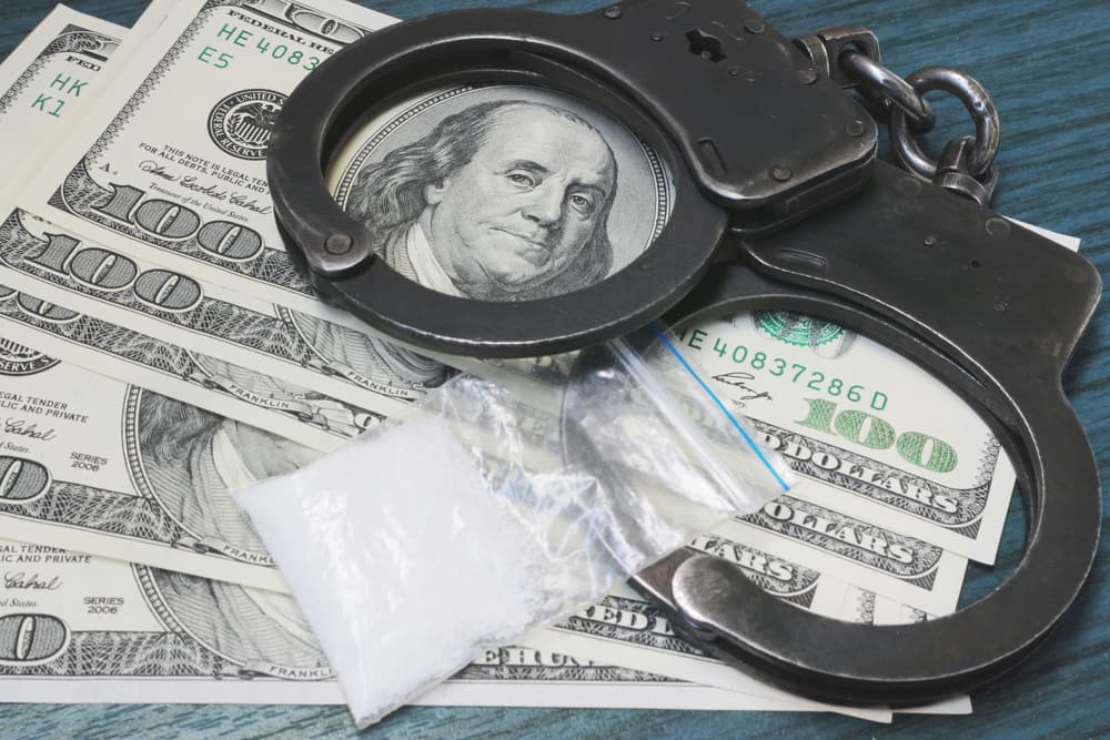 A small bag of white powder, symbolizing narcotic drugs, placed on a table alongside a stack of US dollar bills and a pair of metal handcuffs. The image conveys the concept of legal consequences and punishment for drug possession.